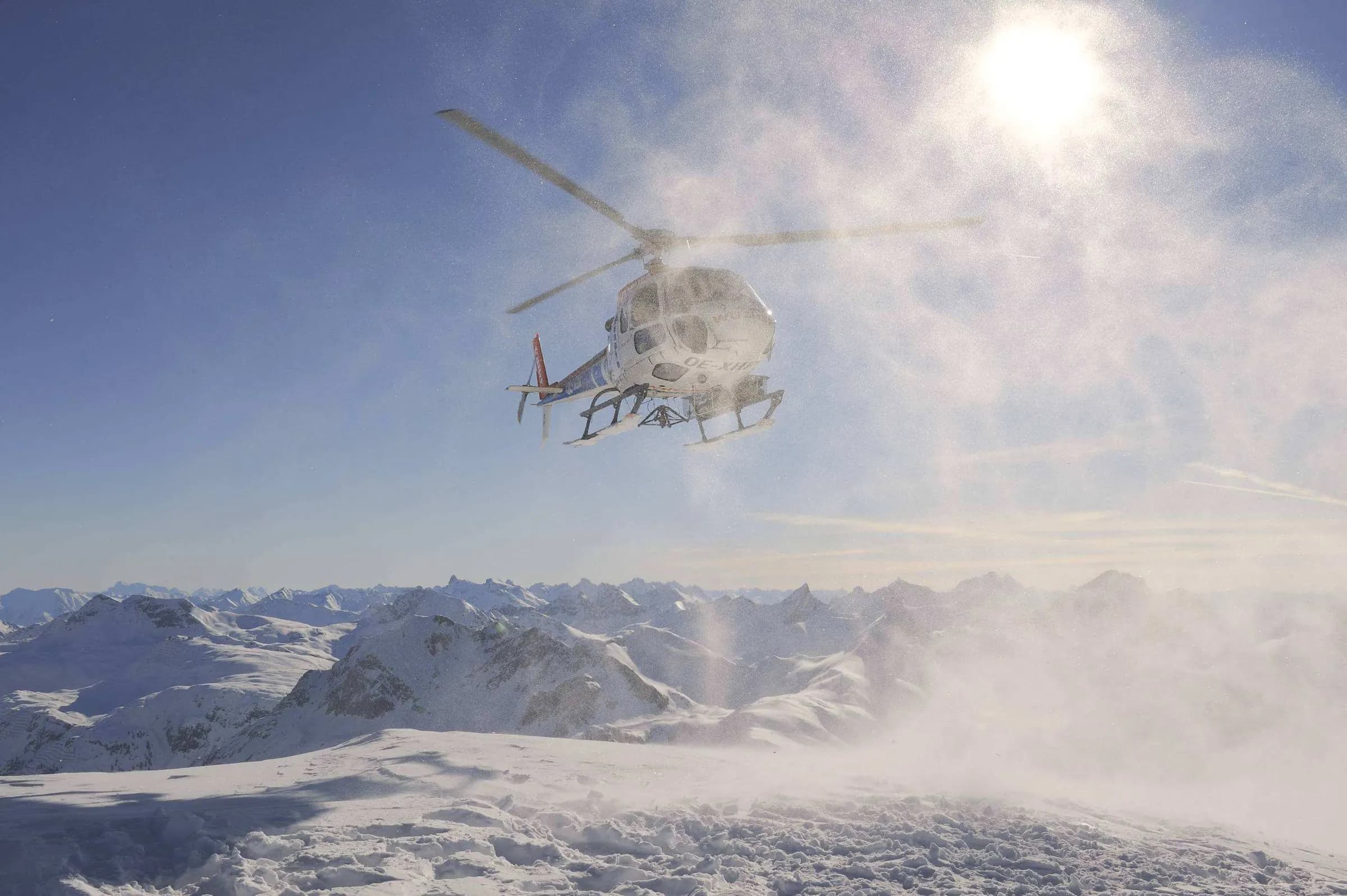 Helikopter in tegenlicht boven de toppen, met opwaaiende sneeuw