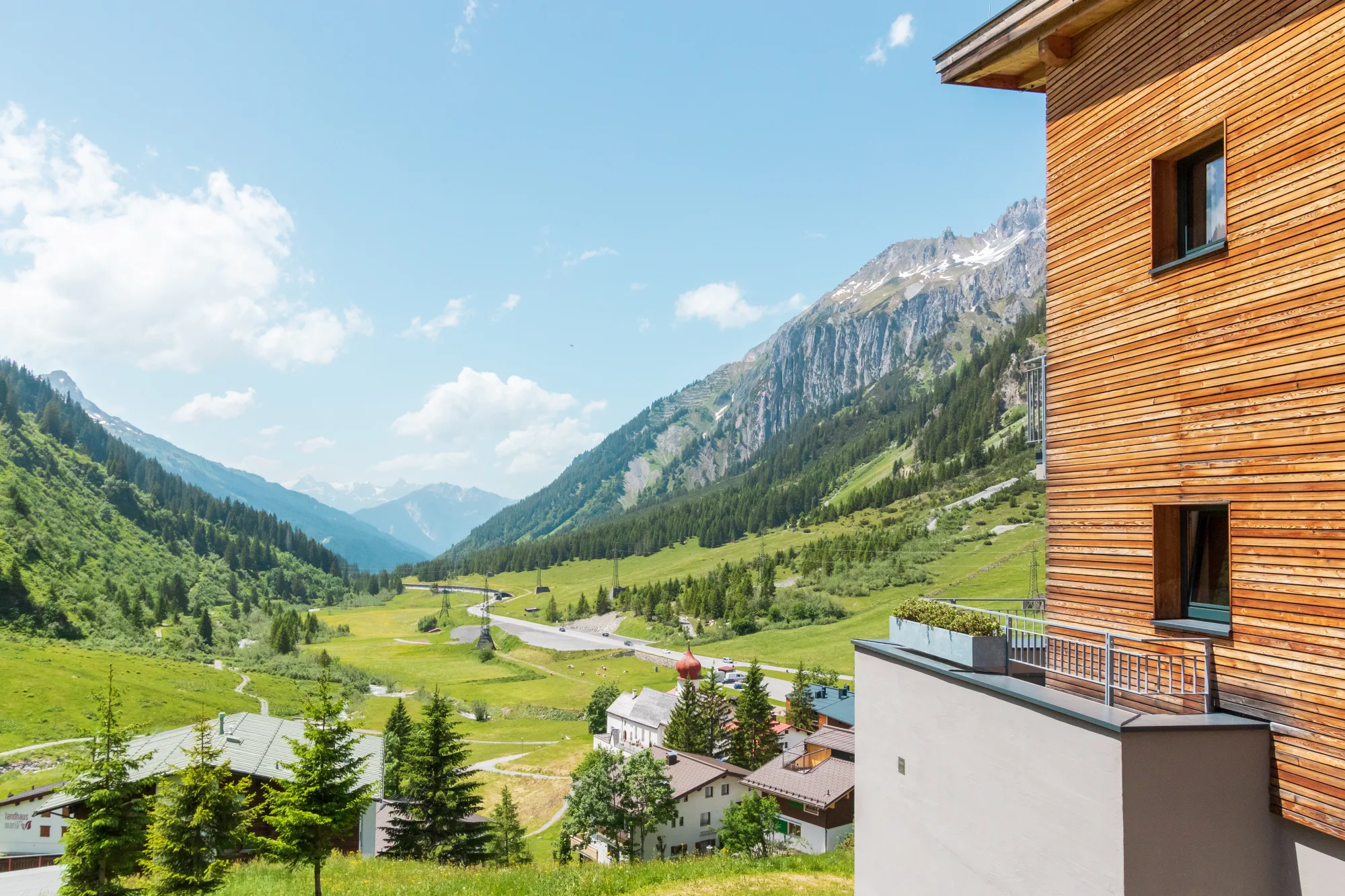 Panoramic summer view from Chalet Mondschein in Stuben am Arlberg with green alpine meadows, mountain peaks and the Arlberg Pass road behind