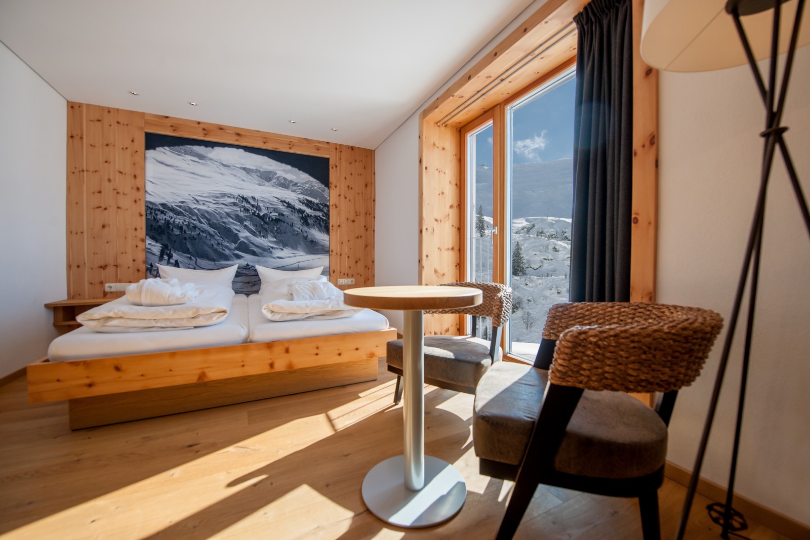 Bright Comfort Double Room at Ski-Hotel Mondschein in Stuben am Arlberg, Austria – featuring a double bed with pine wood frame, panoramic mountain view, historic alpine photo mural, and a cozy sitting area with woven armchairs