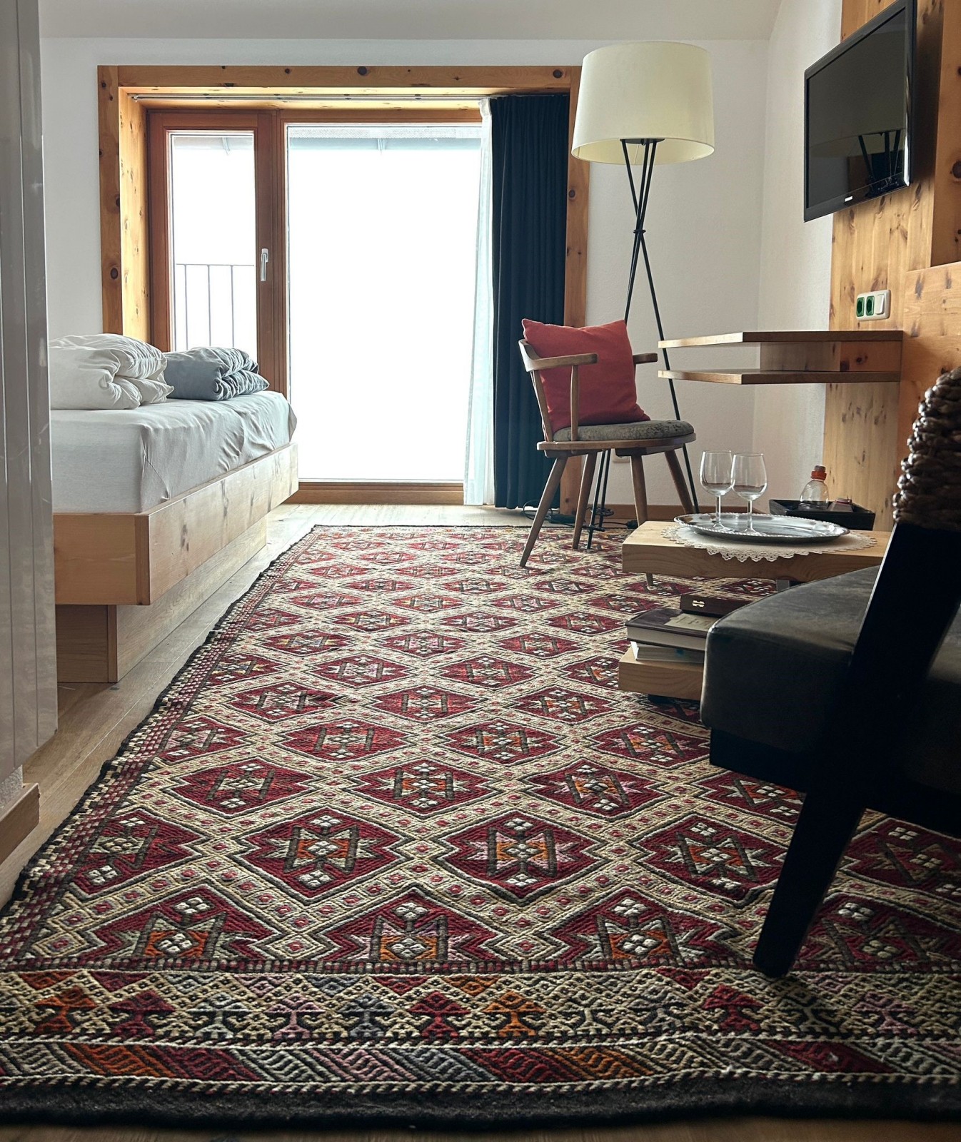 Sitting area of the Comfort Double Room at Ski-Hotel Mondschein in Stuben am Arlberg, Austria – with a red patterned carpet, lounge chair, work desk, TV, and large window with mountain light