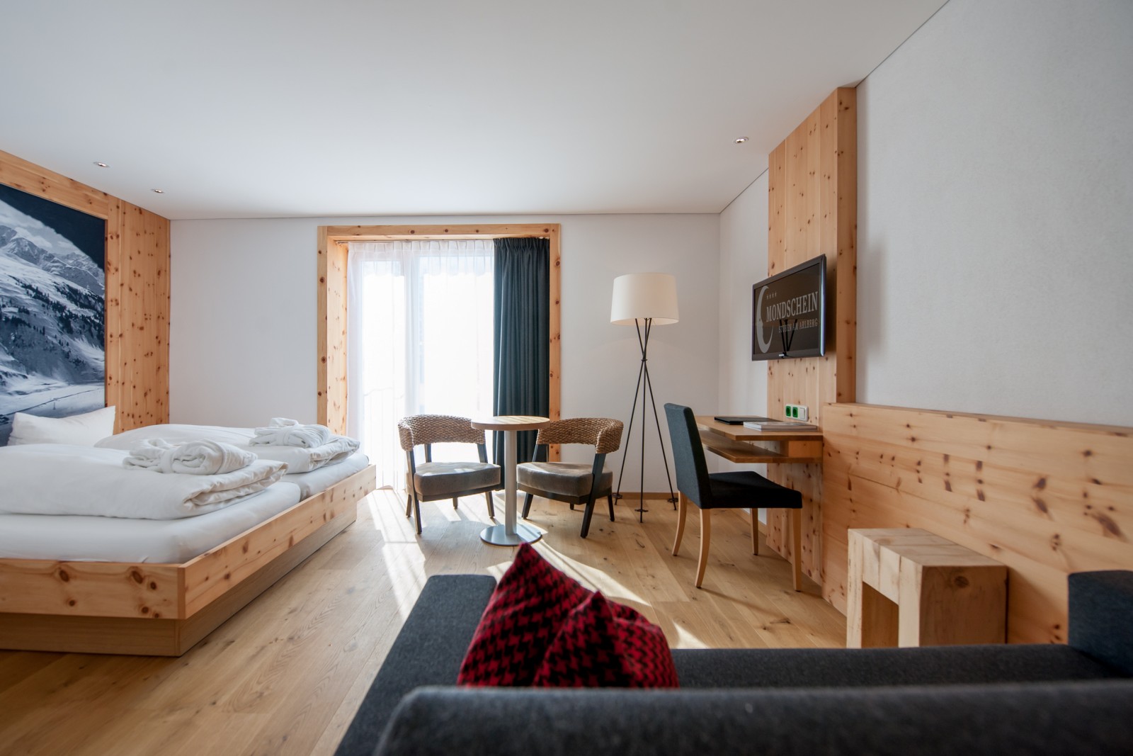 Comfort Triple Room at Ski-Hotel Mondschein in Stuben am Arlberg, Austria – double room with sofa bed, pine wood interior, mountain photo mural, floor-to-ceiling windows, writing desk, and natural wooden floor