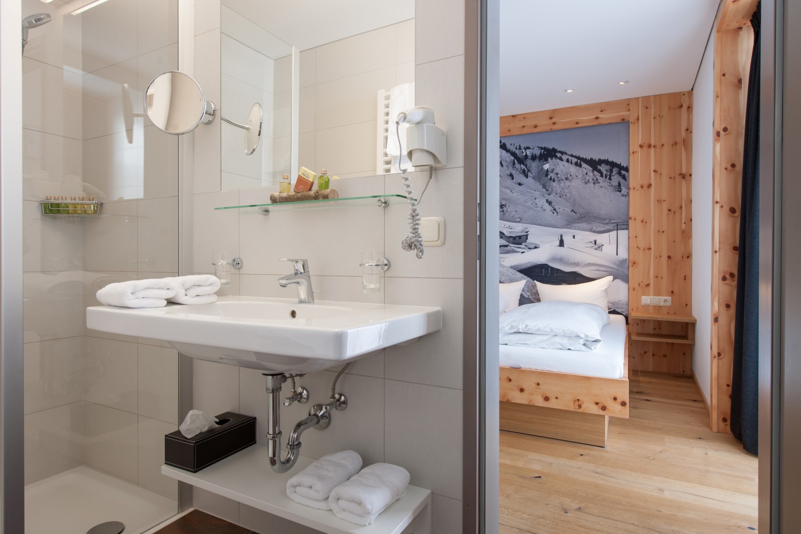 Modern bathroom with walk-in shower, sink, mirror, and hairdryer at Ski-Hotel Mondschein, connected via door to the second bedroom of the Family Mondschein Suite with pine wood interior and alpine photography