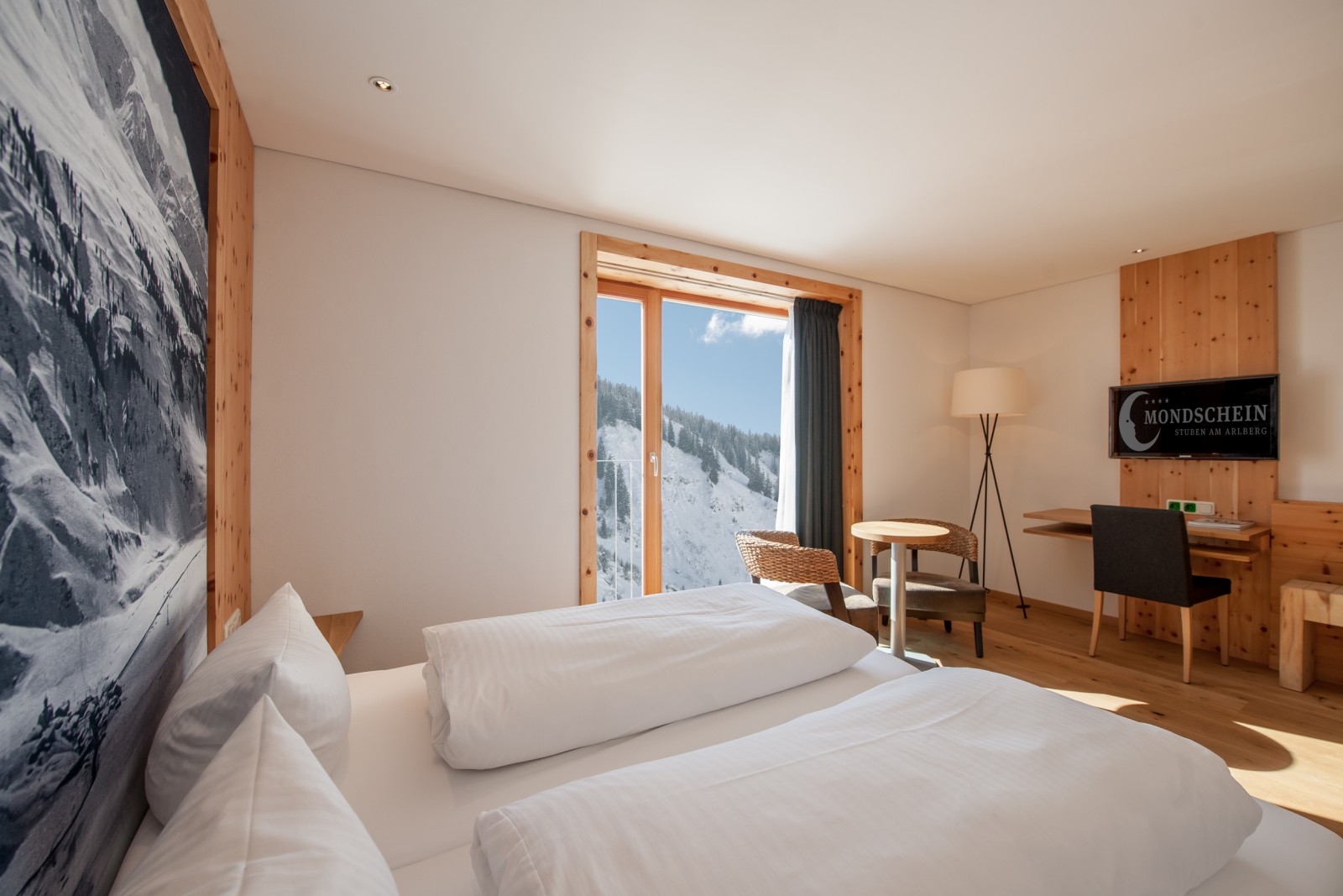 Panorama Double Room at Ski-Hotel Mondschein in Stuben am Arlberg, Austria – featuring a double bed, flat-screen TV, natural pine wood interiors, writing desk, woven chairs, and mountain views through panoramic windows