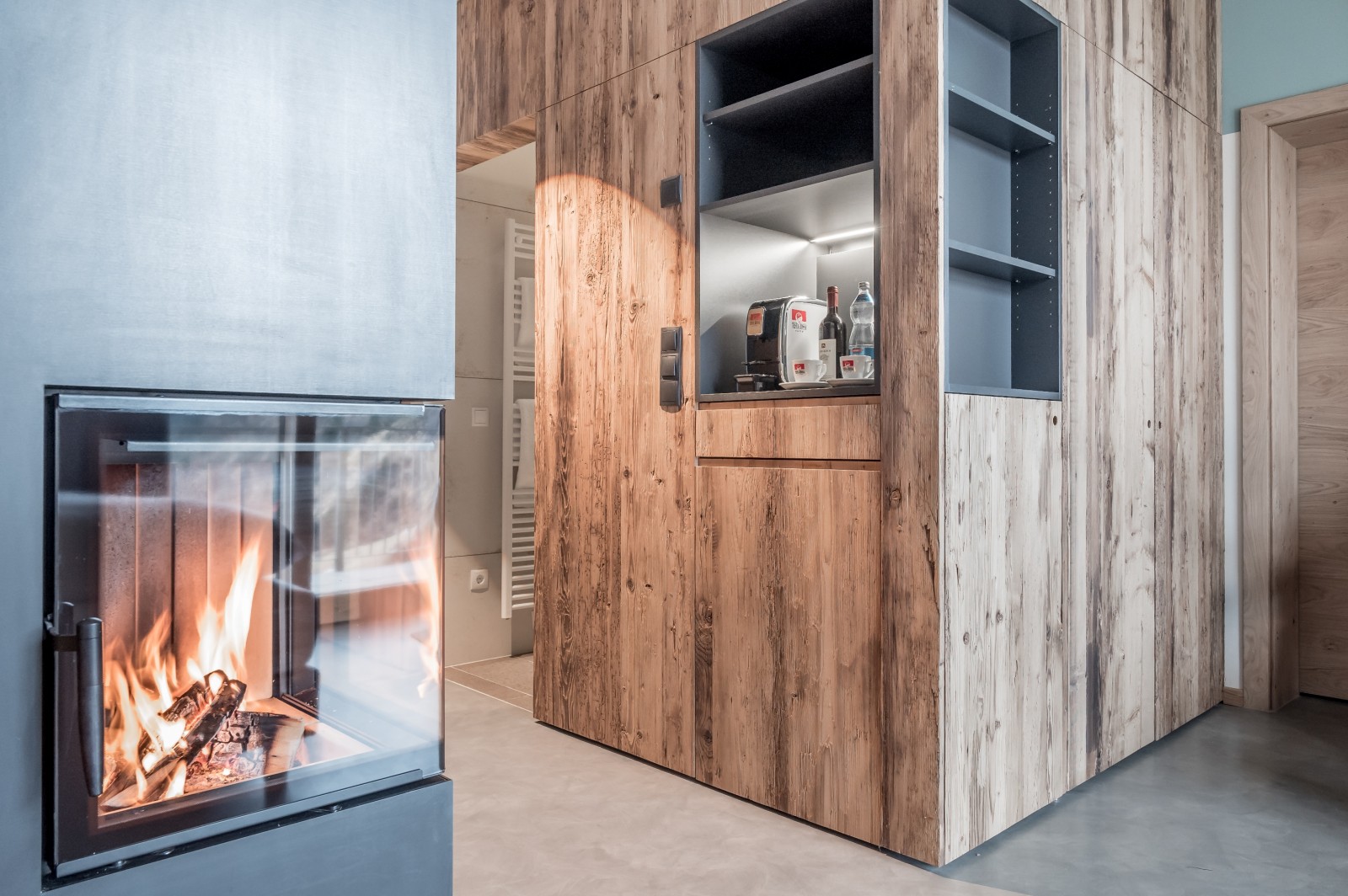 Cozy fireplace and wood-accented coffee corner in the Private SPA Double Deluxe at Chalet Mondschein in Stuben am Arlberg, Austria – featuring a built-in coffee station, open shelving, and modern alpine design