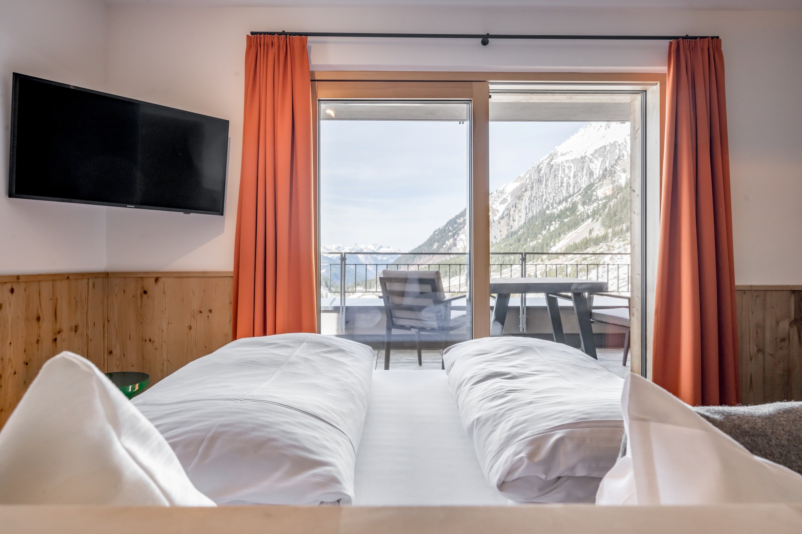 View from the pine wood bed in the Private SPA Double Deluxe at Chalet Mondschein in Stuben am Arlberg, Austria – with balcony, orange curtains, flat-screen TV, and panoramic view of the snow-covered Alps