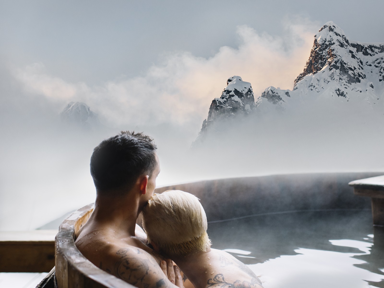 A couple relaxing in a steaming outdoor hot tub at Chalet Mondschein in Stuben am Arlberg, Austria, with panoramic views of misty, snow-covered alpine peaks at sunset