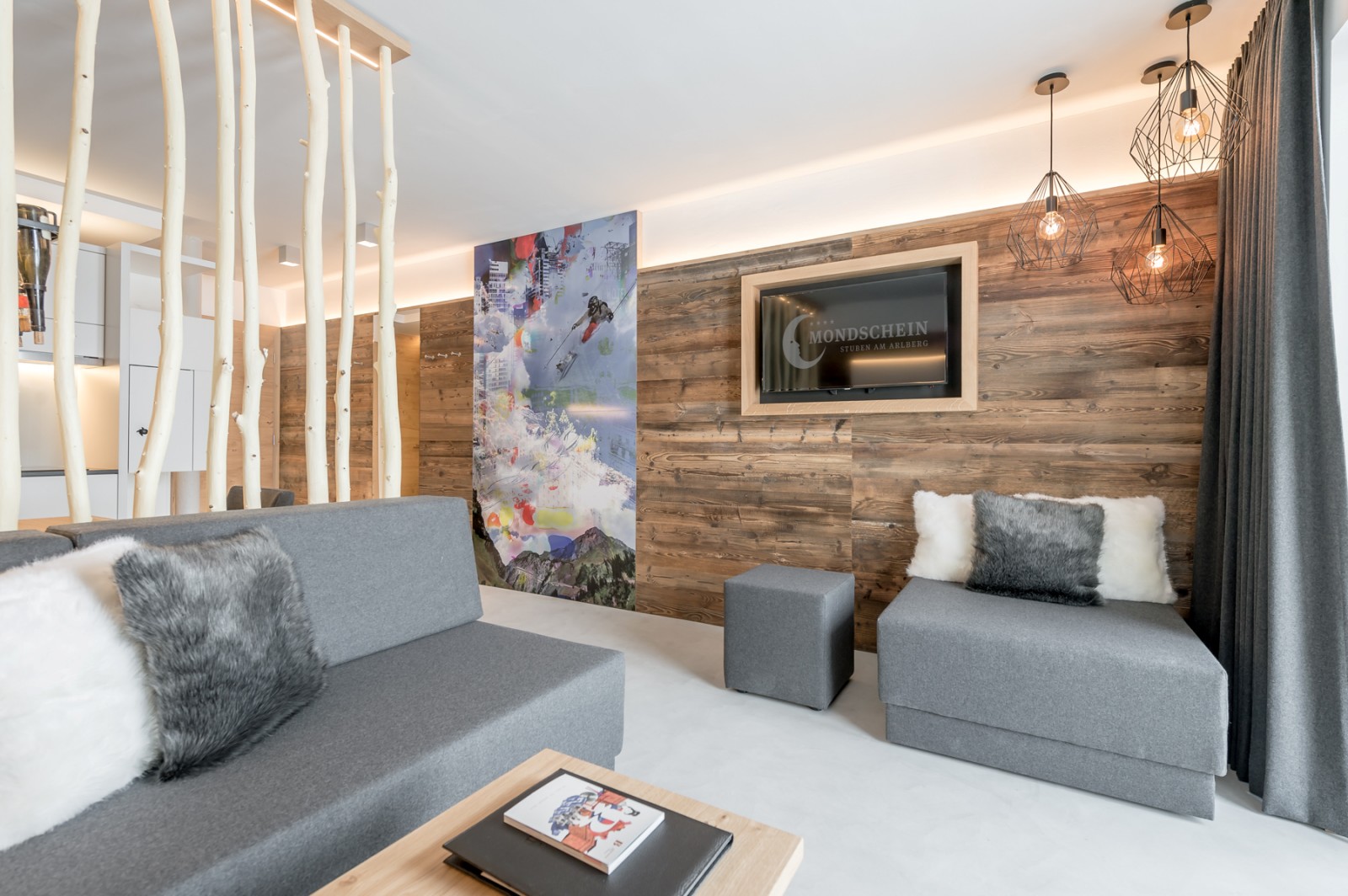 Living room of the Private SPA Suite Premium at Chalet Mondschein in Stuben am Arlberg, Austria – alpine-modern interior with cozy daybeds, faux fur pillows, custom wood paneling, open partition of natural birch wood trunks, flat-screen TV, and design lighting