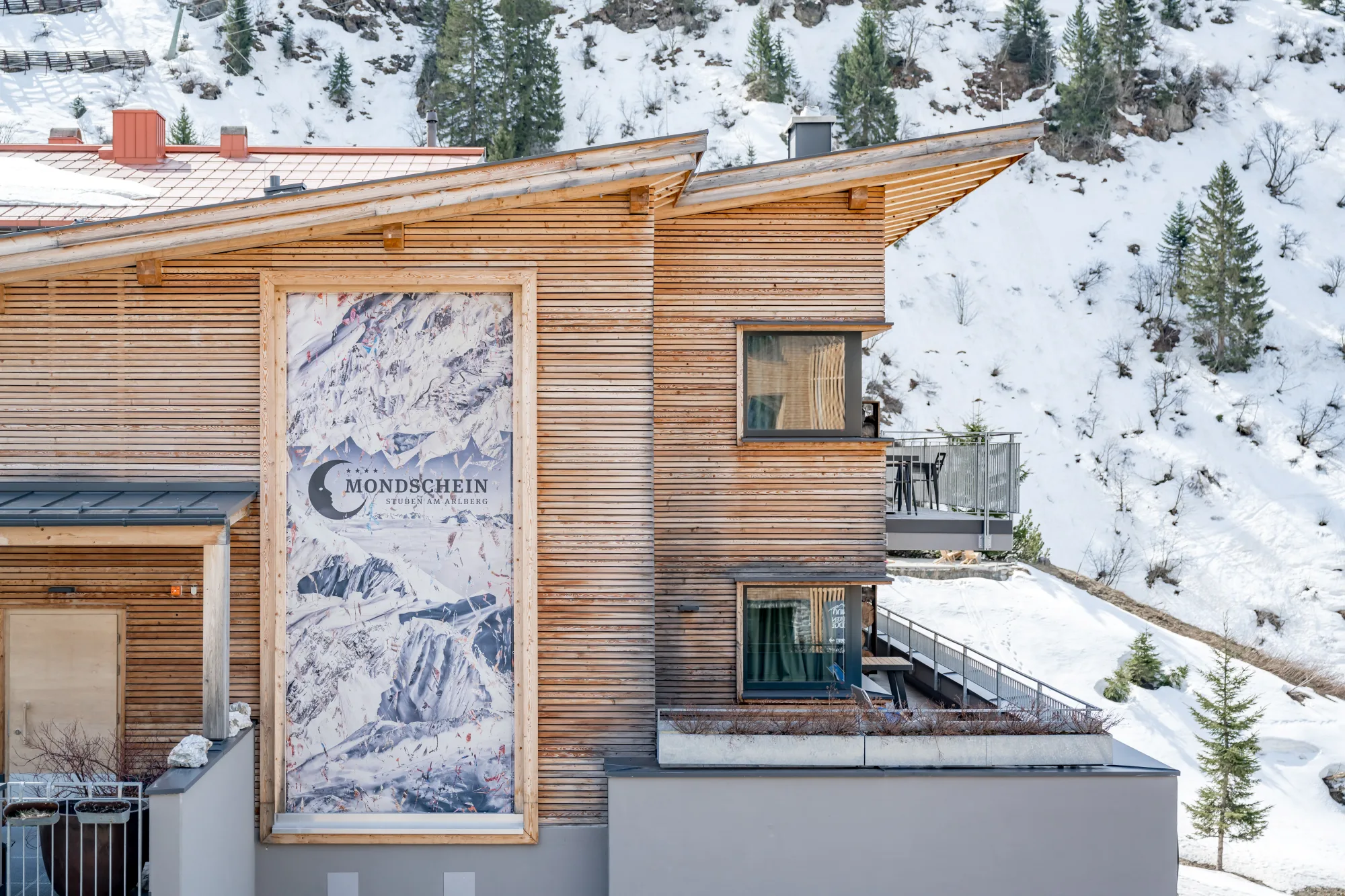 Exterior view of Chalet Mondschein in Stuben am Arlberg, Austria – featuring natural timber cladding, alpine architecture, balconies with panoramic views, and a large mural set against a snowy mountain backdrop