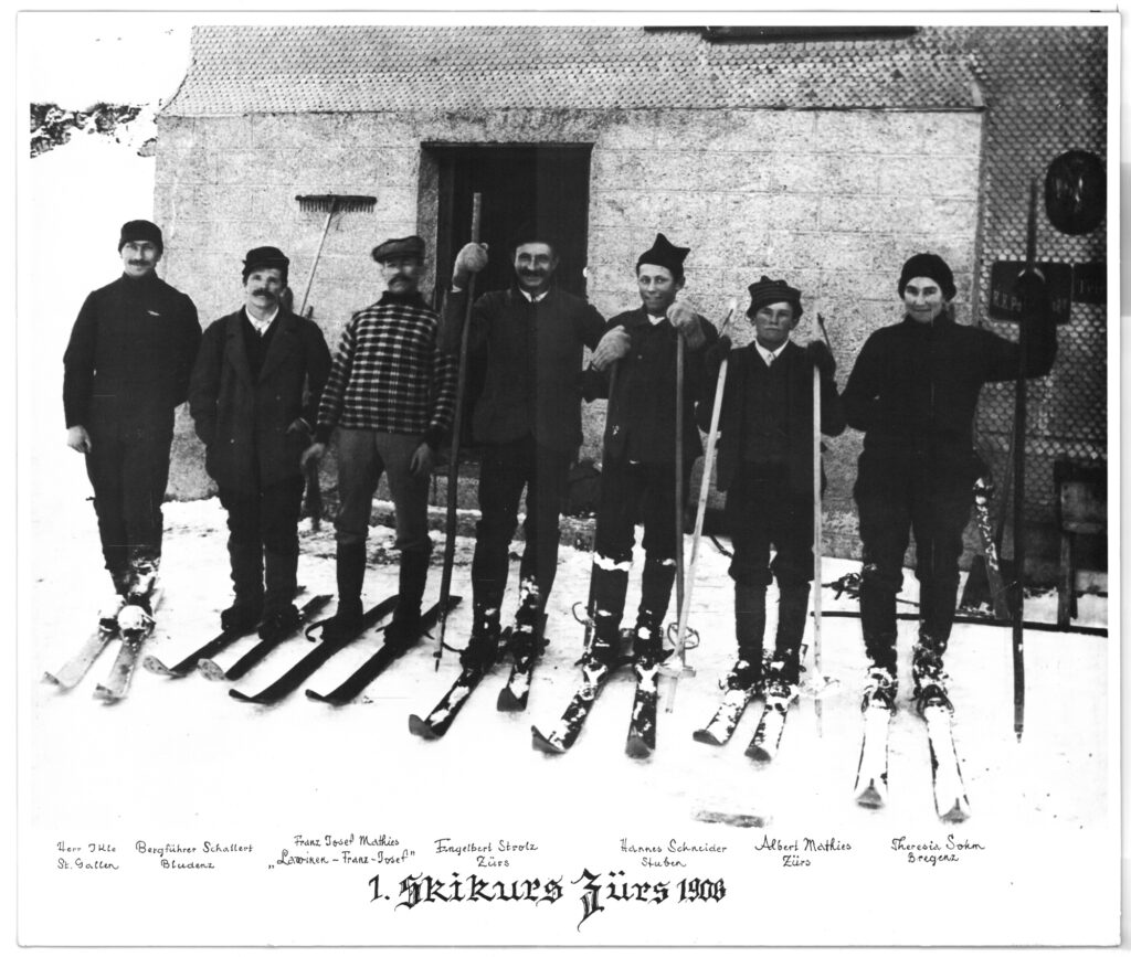 Historical black-and-white photo of the first ski course in Zürs, showing a group of skiers and an instructor on a snow-covered slope in the Austrian Alps