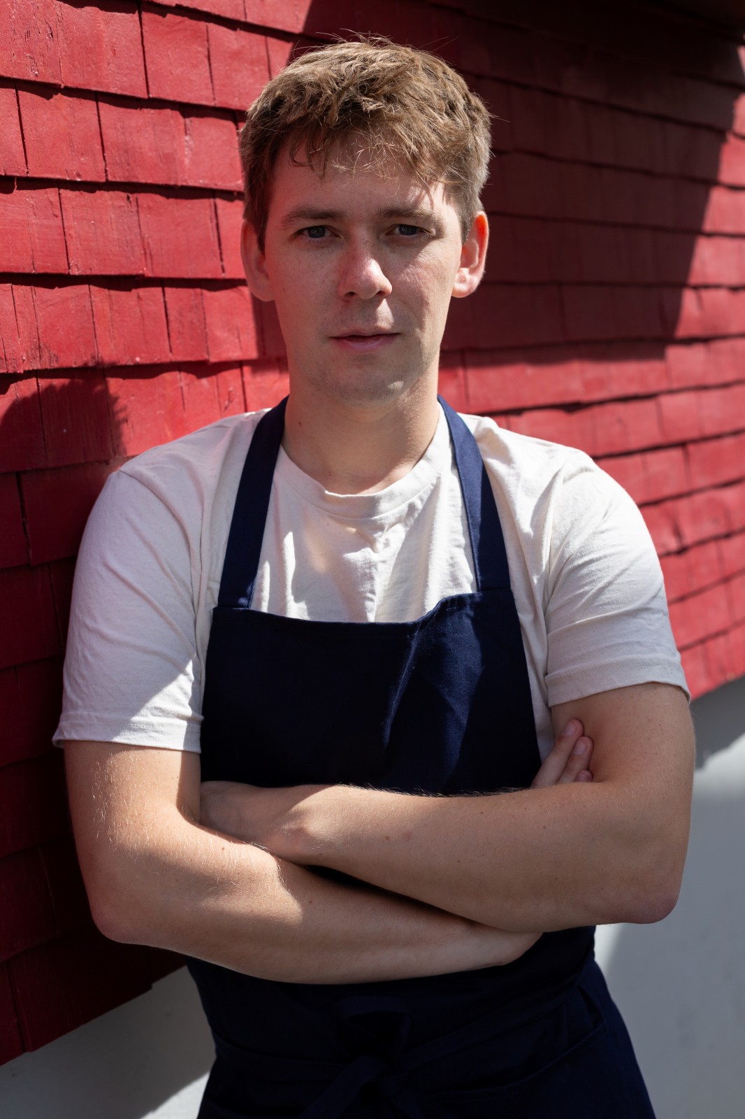Chef Joshua Leise in a white shirt and navy apron, standing in front of a red wooden wall, arms crossed, natural sunlight