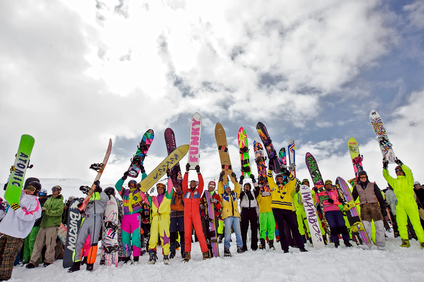 Colorfully dressed snowboarders and skiers raising their vintage boards during the Longboard Classics event at Arlberg under a cloudy winter sky