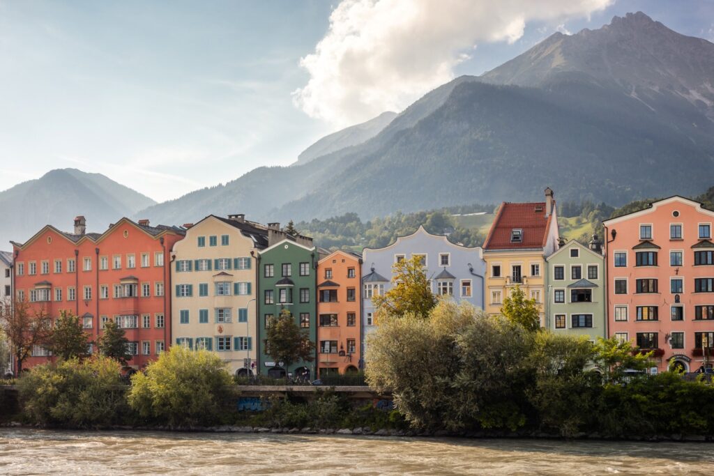 Row of colorful historic townhouses along the Inn River in Innsbruck, Austria, framed by lush trees and alpine mountains in the summer afternoon light