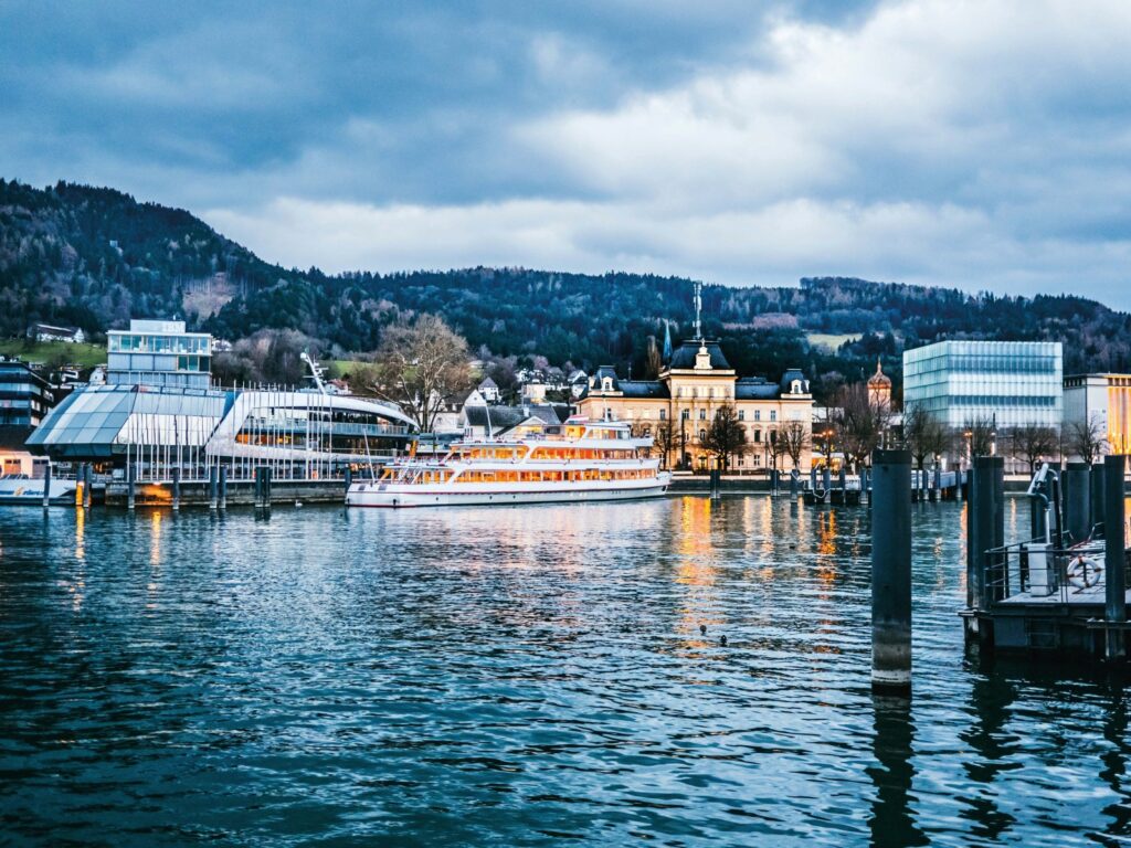 Illuminated boats and cultural buildings along the harbour promenade in Bregenz, Austria, at dusk with Lake Constance in the foreground and forested hills in the background