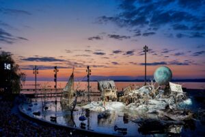 Stunning open-air stage set of “Der Freischütz” at the Bregenz Festival 2023, with dramatic wooden houses, a tilted church tower, and a moon sculpture rising over Lake Constance at sunset
