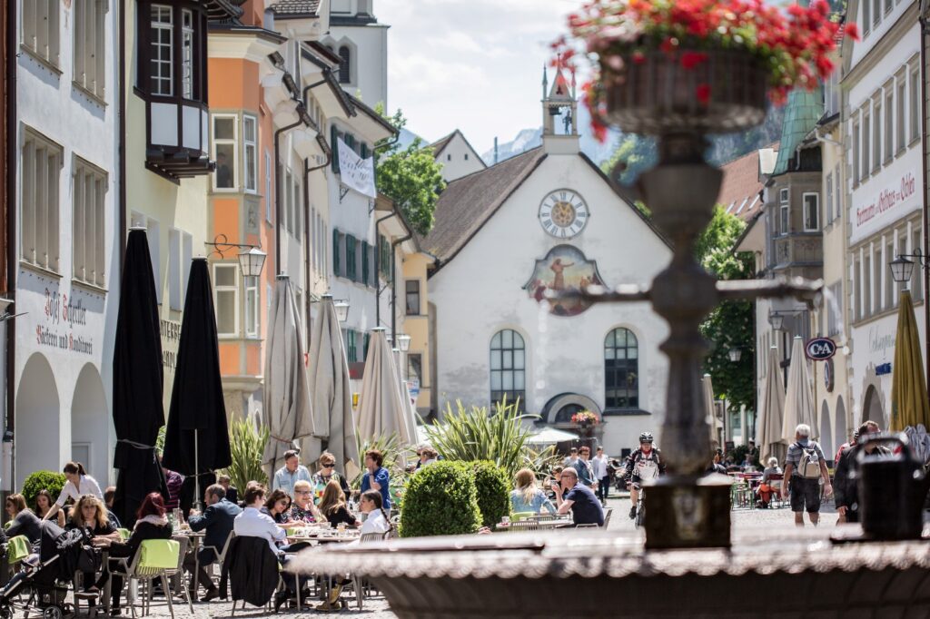 People enjoying outdoor cafés on Marktgasse in Feldkirch, Austria, with colorful historic townhouses and the old city church in the background on a sunny summer day
