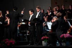 Male soloists and conductor performing with orchestra and choir at the Lech Classic Festival 2023 in Austria, dressed in formal evening attire, surrounded by musicians and flowers on stage